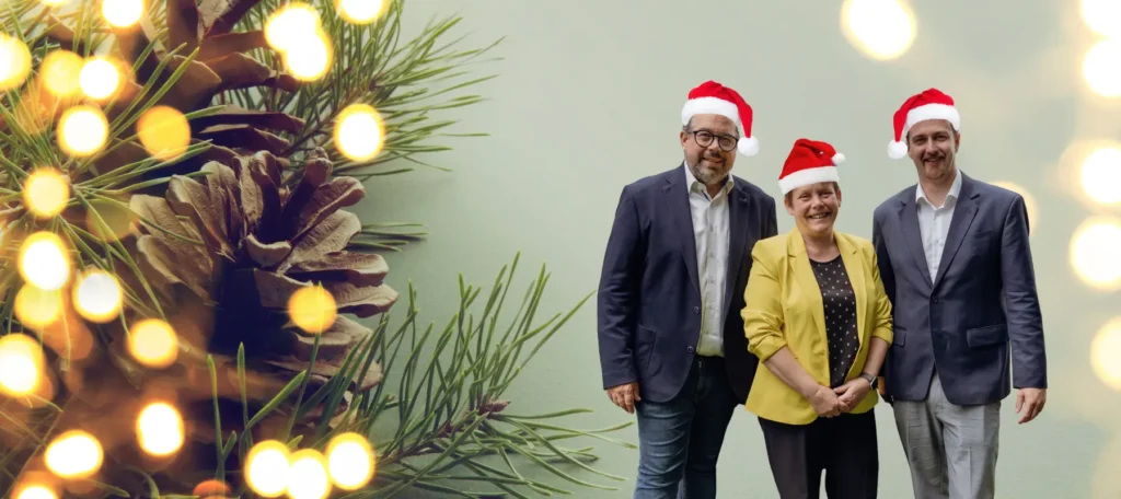 Zwei Männer und eine Frau mit Weihnachtsmützen vor einem weihnachtlichen Hintergrund